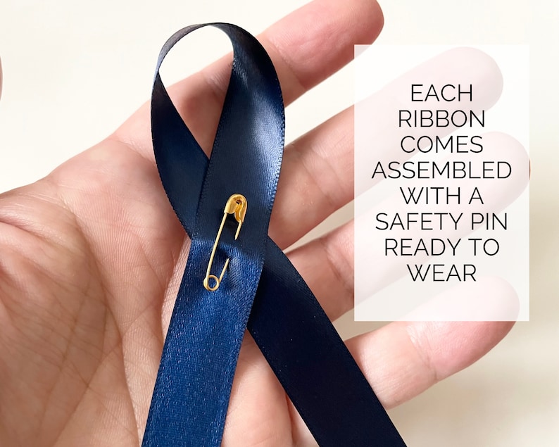 May include: A dark blue ribbon with a gold safety pin attached. The ribbon is tied in a loop and is ready to wear. The text "EACH RIBBON COMES ASSEMBLED WITH A SAFETY PIN READY TO WEAR" is visible in the background.