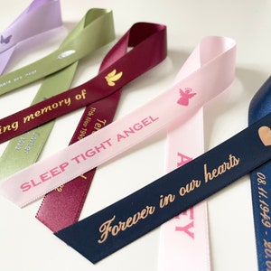 May include: Five satin ribbons with different colors and messages printed in gold and pink. The messages include "In loving memory of", "Sleep Tight Angel", "Forever in our hearts", and "Lennon Rhe 08.11.1949 - 20.08".