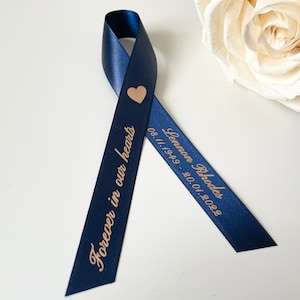May include: A navy blue ribbon with a gold heart and the text "Lennon Rhodes 08.11.1949 - 20.01.2022" and "Forever in our hearts" printed on it.