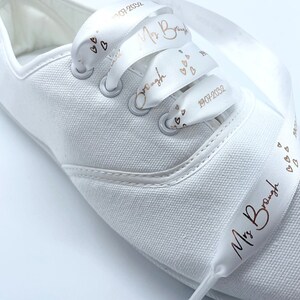 May include: White canvas sneaker with satin ribbon laces. The ribbon is printed with the text "Mrs. Brough", hearts, and the date "19.07.2032" in a copper color. The shoe has silver eyelets.