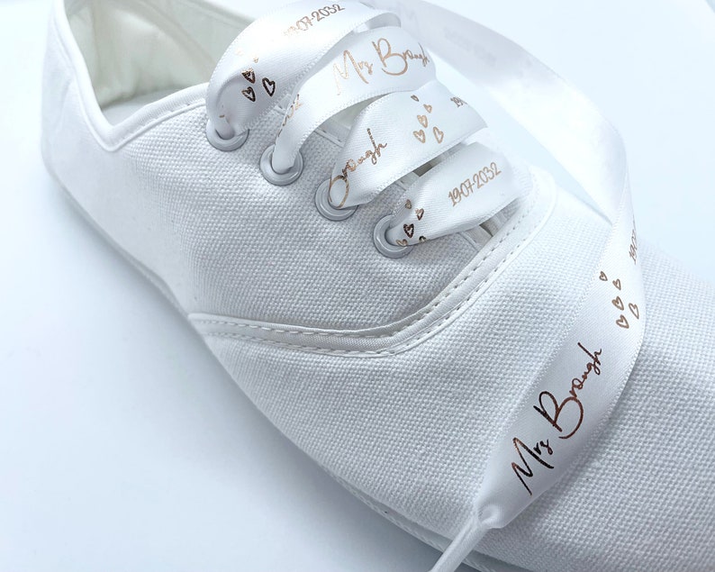 May include: White canvas sneaker with satin ribbon laces. The ribbon is printed with the text "Mrs. Brough", hearts, and the date "19.07.2032" in a copper color. The shoe has silver eyelets.