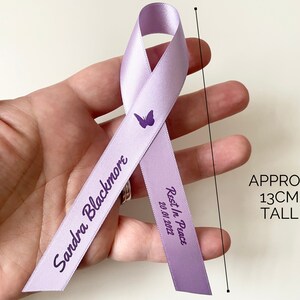 May include: A lavender ribbon with a butterfly design and the text "Sandra Blackmore Rest In Peace 20.01.2022". The ribbon is approximately 13 inches tall.