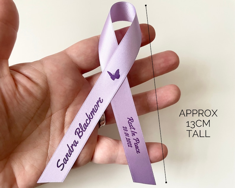 May include: A lavender ribbon with a butterfly design and the text "Sandra Blackmore Rest In Peace 20.01.2022". The ribbon is approximately 13 inches tall.