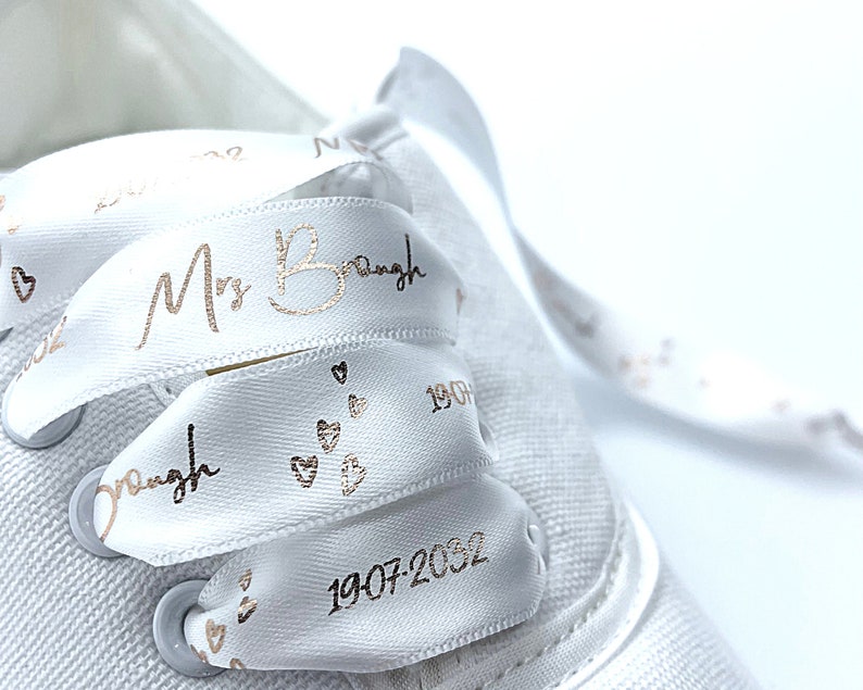 May include: White satin shoelaces with rose gold text and heart accents. The text reads "Mrs. Braugh" and a date. The laces are threaded through a white canvas shoe. A close-up shot highlights the details of the personalized laces.