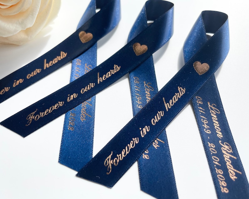 May include: Three navy blue satin ribbons with gold hearts and the text "Forever in our hearts" printed on them. The ribbons are tied together in a knot.