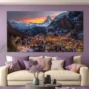 May include: A large canvas print depicting a mountain village at sunset. Snow-covered mountains surround the town, with the Matterhorn peak visible. The sky is a mix of orange and yellow, and the village lights are illuminated.