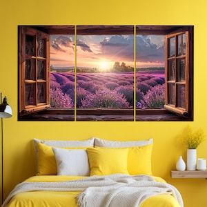 May include: A three-panel canvas print featuring a view of a lavender field through an open window. The field is bathed in the golden light of a setting sun, with a blue sky and fluffy clouds above. The window frame is brown wood, and the window panes are clear glass.
