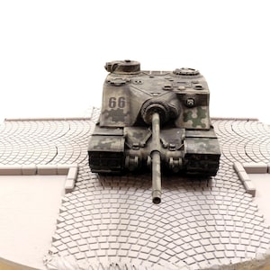 Op de afbeelding: Een model van een militaire tank met camouflageverf en het nummer 66 aan de zijkant. De tank staat op een grijze grindsteenbasis.