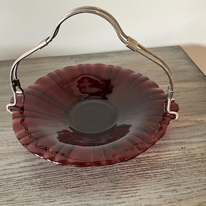 May include: A vintage burgundy glass serving basket with a silver-toned handle. The basket has a scalloped rim and a deep, rounded bowl. The handle is arched and attached to the sides of the basket. A decorative piece for serving or display.