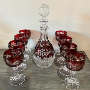 Puede incluir: Un decantador de cristal con tapón a juego y seis copas de tallo, todos con una capa de color rojo intenso y grabados de motivos de uvas y vides. El decantador tiene una base transparente con cortes verticales y las copas tienen tallos claros.