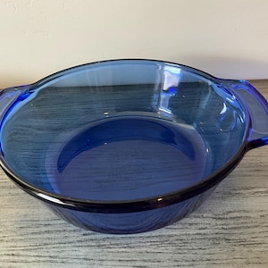 Puede incluir: Un cuenco de vidrio azul cobalto vintage con dos asas integradas. El cuenco tiene forma redondeada y un borde oscuro. El vidrio es transparente, permitiendo el paso de la luz. Este artículo de cocina es adecuado para servir o hornear.