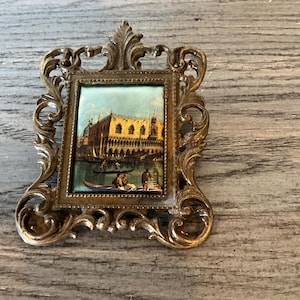 May include: Ornate, vintage-style picture frame with a rectangular painting of a Venetian scene. The frame is gold-toned with intricate scrollwork. The painting depicts a canal with gondolas and buildings.