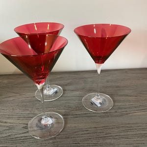 Könnte beinhalten: Drei rote Martinigläser mit klaren Stielen und Böden. Die Gläser haben eine klassische dreieckige Schalenform und sind aus Glas. Die Gläser sind auf einer Holzoberfläche angeordnet.