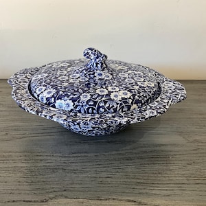 May include: A round ceramic serving dish with a matching lid, featuring a dark blue and white floral pattern throughout. The lid has a small, sculpted handle at the center, and the dish has a wide, scalloped rim with a continuous botanical design.