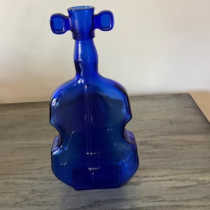 May include: A deep blue glass bottle in the shape of a violin. The bottle features a long neck with two rounded elements at the top, and the body of the violin has etched lines. It is placed on a grey wooden surface.