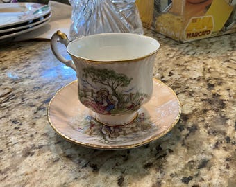 May include: A vintage teacup and saucer set. The cup and saucer are light pink with gold trim. The cup features a pastoral scene with a couple under a tree. The saucer has a matching design. A classic addition to any tea set collection.