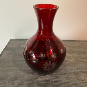 May include: A deep red glass vase with a bulbous body and a flared rim. The surface of the vase features etched floral patterns and circular motifs that reveal clear glass beneath the red color, creating a contrast in texture and light.
