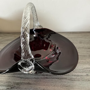 May include: A dark burgundy glass basket with a clear, twisted glass handle. The basket has a wide, shallow bowl shape with a flared rim. The handle is arched and attached to the bowl. The glass has a glossy finish.