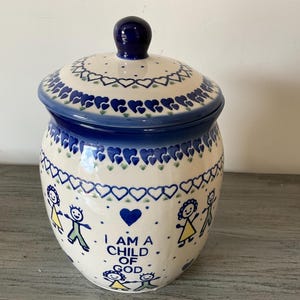 May include: A ceramic jar with a lid, featuring a blue and white heart pattern and the text "I AM A CHILD OF GOD." The jar has a dark blue knob on top and is decorated with stylized figures.