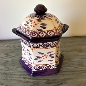 Peut inclure: Un pot en céramique avec un couvercle, de forme hexagonale et orné d'un motif floral décoratif. Le pot a une base blanche avec des accents violets et rouges, et un bouton marron foncé sur le couvercle. Le pot est posé sur une surface en bois.