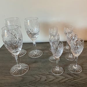May include: A collection of clear crystal wine glasses and cordial glasses. The glasses have etched floral patterns and starburst bases. The stemware is arranged on a dark wooden surface.