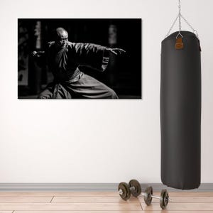 May include: A black and white photograph of a man in a martial arts pose. He is wearing a traditional robe and has a serious expression on his face. The photo is hanging on a white wall next to a black punching bag and a set of dumbbells.