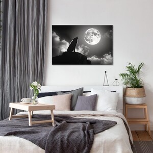 May include: Black and white wall art print of a wolf howling at the moon. The artwork depicts a silhouette of a wolf on a rocky outcrop, with a full moon in a cloudy night sky. The print is displayed above a bed.
