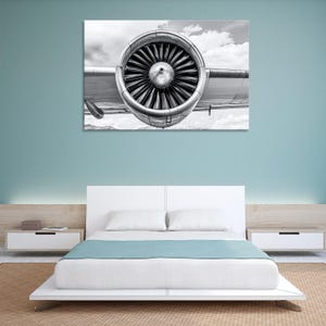 Könnte beinhalten: Schwarz-Weiß-Fotografie eines Flugzeugtriebwerks, die sich auf die Lüfterblätter und das Metallgehäuse konzentriert. Das Bild ist vor einem bewölkten Himmel aufgenommen. Das Kunstwerk wird über einem weißen Bett mit einer blauen Decke gezeigt.