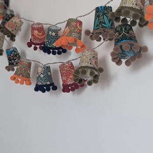 May include: A decorative string of lights with bell-shaped shades. Each shade displays a different floral pattern in various colours, and is trimmed with pom-poms. The lights are strung on a silver wire, creating a colourful and festive display.