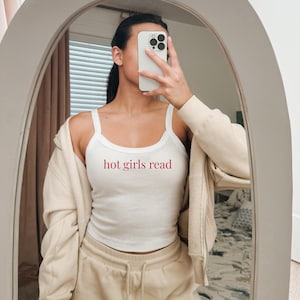 以下が含まれることがあります： 赤い文字で「hot girls read」と書かれた白いタンクトップ。ベージュのジップアップパーカーとベージュのスウェットパンツを着用。鏡は楕円形です。