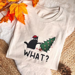 Peut inclure: T-shirt crème avec un chat noir portant un bonnet de Père Noël, interagissant avec un sapin de Noël. Le mot « WHAT ? » est imprimé en dessous. Le t-shirt est présenté sur une surface blanche et duveteuse avec des feuilles d'automne.
