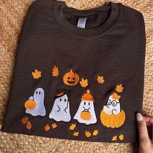 Pode incluir: Um moletom marrom com bordados temáticos de Halloween. O design inclui quatro figuras de fantasmas, uma abóbora esculpida e folhas de outono em laranja e amarelo. O moletom é feito de um material macio.