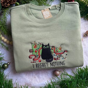 Puede incluir: Sudadera verde salvia con un diseño bordado de un gato negro frente a un árbol de Navidad con adornos y luces rojas. El texto "I REGRET NOTHING" está debajo del gato.
