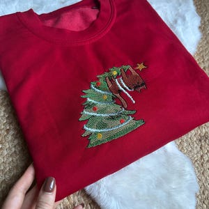 Puede incluir: Una sudadera roja de cuello redondo con un diseño de árbol de Navidad bordado. El árbol es verde con adornos rojos y amarillos, una estrella dorada y una cabeza de caballo marrón. La sudadera es de material suave.
