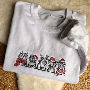 Puede incluir: Sudadera blanca con un bordado de cinco gatos con accesorios festivos. Los gatos llevan una bufanda roja, gorros de Papá Noel y cuernos de reno. La sudadera está hecha de un material suave.