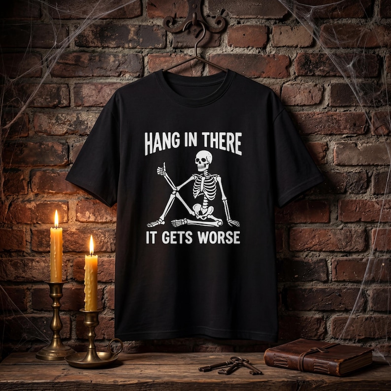 Może przedstawiać: Czarny t-shirt z kr&oacute;tkim rękawem z białą grafiką przedstawiającą siedzący szkielet pokazujący kciuk w g&oacute;rę. Tekst nad szkieletem brzmi HANG IN THERE, a tekst poniżej brzmi IT GETS WORSE, napisany pogrubioną, białą czcionką.