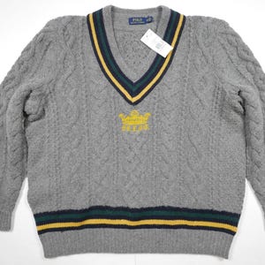 May include: Gray cable knit sweater with a v-neck and a yellow embroidered crown with the word "Polo" on it. The sweater has a navy blue, yellow, and green stripe around the neck and bottom.