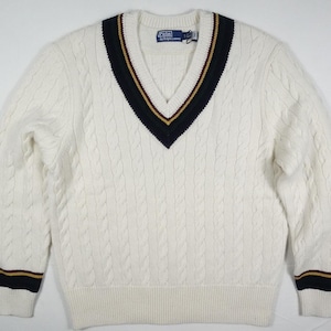 May include: White cable knit sweater with a navy blue and gold trim around the v-neck. The sweater has a Polo Ralph Lauren label.