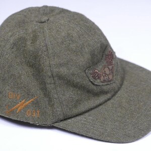 May include: A green wool baseball cap with a brown embroidered patch on the side. The patch features a stylized eagle with wings spread. The cap also has a brown embroidered logo on the front that says "BV 031".