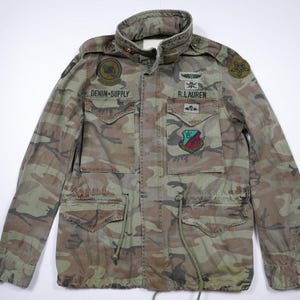 Op de afbeelding: Camouflage jas met meerdere zakken, een opstaande kraag en trekkoorden. De jas heeft patches met de tekst "DENIM SUPPLY" en "R. LAUREN". Het kleurenpalet omvat tinten groen, bruin en beige.