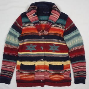 May include: A colorful, patterned cardigan with a shawl collar. The sweater features a mix of red, blue, orange, and teal stripes and geometric designs. It has two front pockets and is fastened with silver buttons.