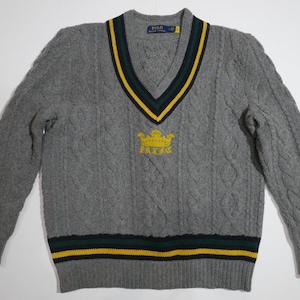 May include: Gray cable knit sweater with a green and yellow striped v-neck collar and a gold crown with the text "PRYDE" embroidered on the front.