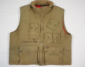 Puede incluir: Un chaleco acolchado de color canela con múltiples bolsillos y un cuello alto. El chaleco tiene un forro rojo y botones dorados. El chaleco es sin mangas y parece estar hecho de un material duradero y resistente al agua.