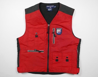 Puede incluir: Chaleco acolchado rojo y negro con cierre de cremallera y múltiples bolsillos. El chaleco tiene un parche con el logotipo de Polo Sport en la parte delantera.