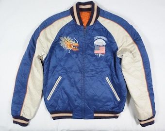 May include: A blue satin bomber jacket with white and orange sleeves. The jacket has a zipper closure and two pockets. The front of the jacket features an embroidered dragon on the left side and a parachute with an American flag on the right side. The text "USAF" is embroidered below the parachute.