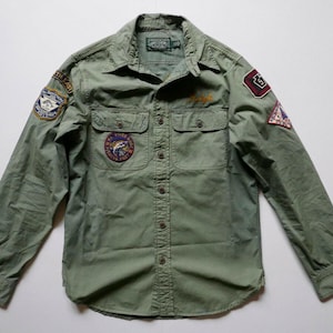 Op de afbeelding: Een groen, lang mouwig overhemd met knopen en meerdere geborduurde patches. De patches hebben verschillende ontwerpen, waaronder een vis, een logo en een nummer.