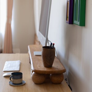 K&ouml;nnte beinhalten: Ein heller Holzschreibtisch mit einem Computermonitor, einem Stifthalter aus Holz und einer blau-grauen Tasse. Auf dem Schreibtisch liegen auch ein kleines Notizbuch und eine Computermaus. Dekorative farbige Rahmen h&auml;ngen an der Wand.