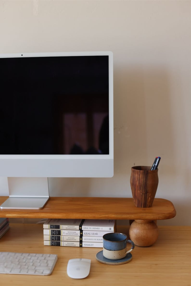 K&ouml;nnte beinhalten: Ein Holzschreibtisch mit einem Computermonitor auf einem Holzst&auml;nder. Eine blau-wei&szlig;e Keramiktasse steht auf einer Untertasse, neben einem Holzstifthalter und einem Stapel B&uuml;cher. Eine Tastatur und eine Maus sind ebenfalls zu sehen.