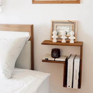 May include: A wall-mounted wooden shelf with an L-shaped design featuring two horizontal surfaces. The top surface holds three white busts and a framed picture, while the lower surface holds a brown candle and two books titled The Kinfolk Table and This Is Home.