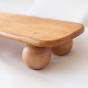 K&ouml;nnte beinhalten: Ein helles Holzserviertablett mit abgerundeten Kanten, das von drei kugelf&ouml;rmigen Holzf&uuml;&szlig;en getragen wird. Das Tablett hat eine nat&uuml;rliche Holzmaserung und eine glatte Oberfl&auml;che, ideal f&uuml;r die Pr&auml;sentation von Speisen oder Dekorationen.